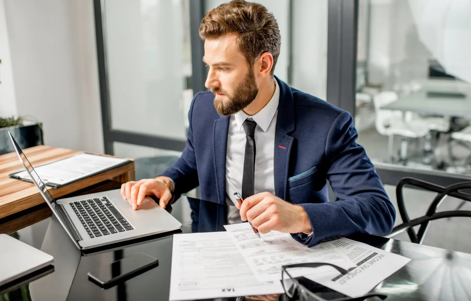 A bearded man in a navy blue suit, white shirt, and black tie sits at a black glass desk, typing on a silver laptop with one