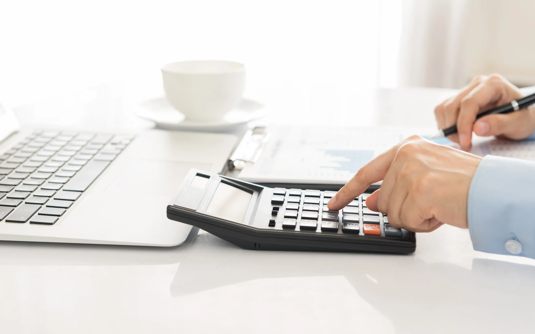 Person using calculator at desk with laptop, coffee cup, and financial documents.