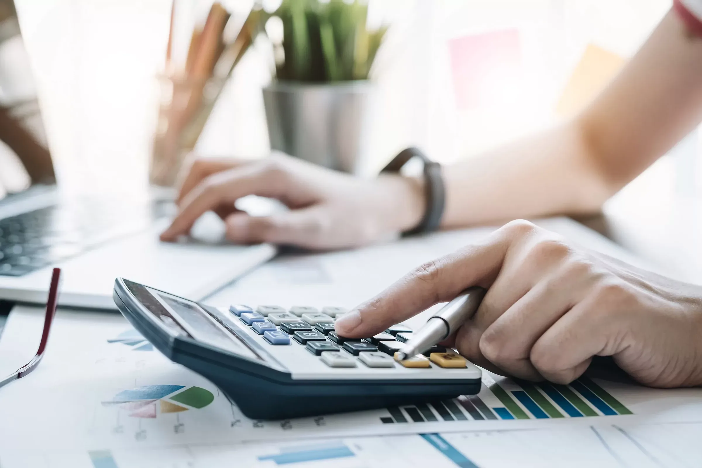 Person using calculator while reviewing financial charts and data on desk with laptop.