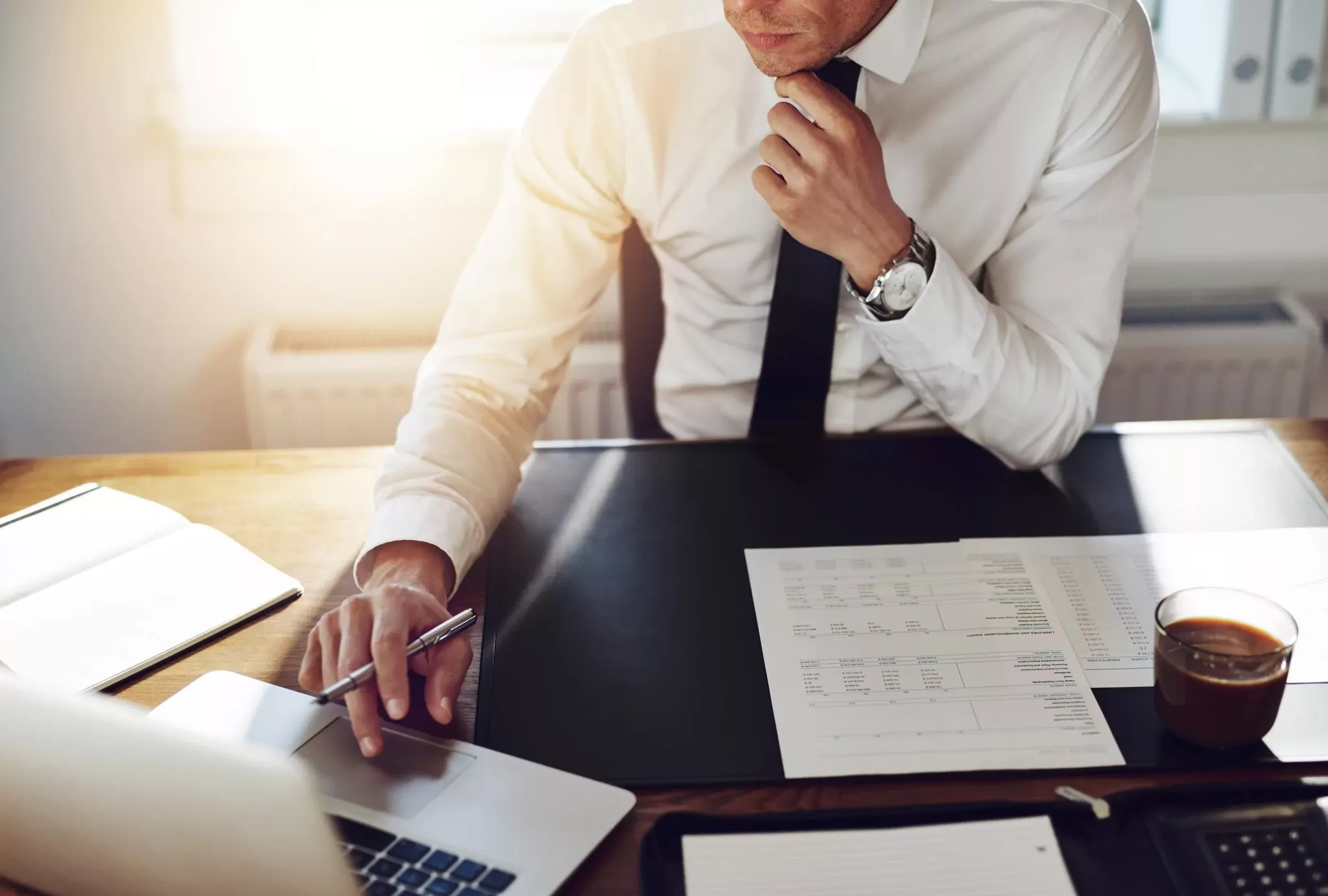 Professional man in white shirt and tie reviewing financial documents and spreadsheets at desk with laptop.