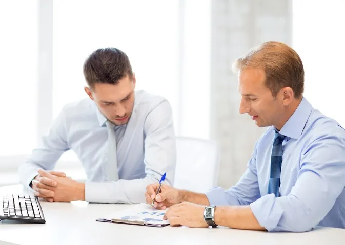 Two businessmen in blue shirts reviewing and signing documents at a desk.