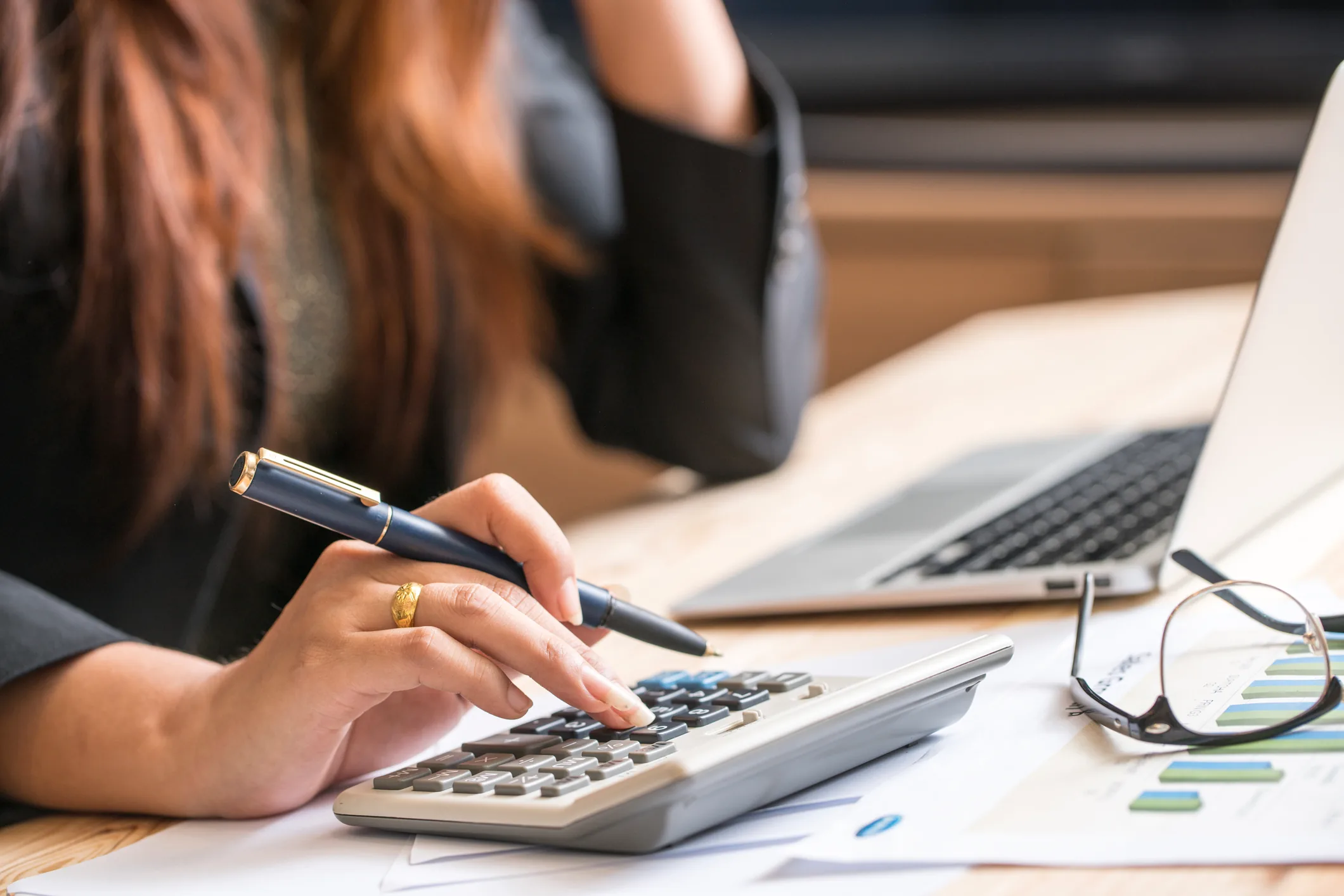 Woman using calculator and pen while reviewing financial documents and laptop at desk.